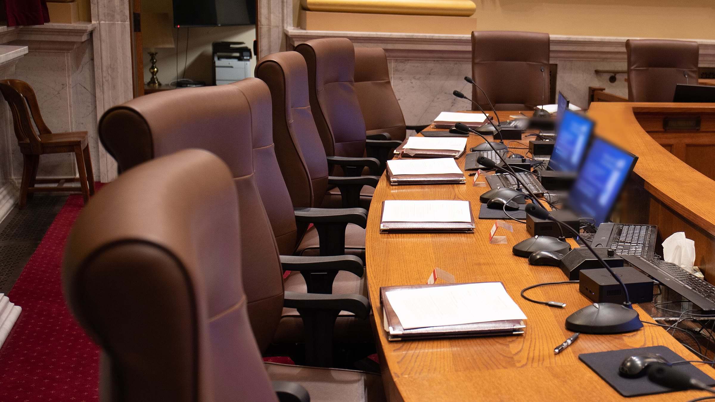 A council chamber's dais: leather chairs, microphones and meeting packets along a curved wooden desk.